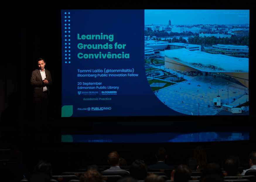 A partially-obscured man stands onstage in front of a screen with text reading "Learning Grounds for Convivência"