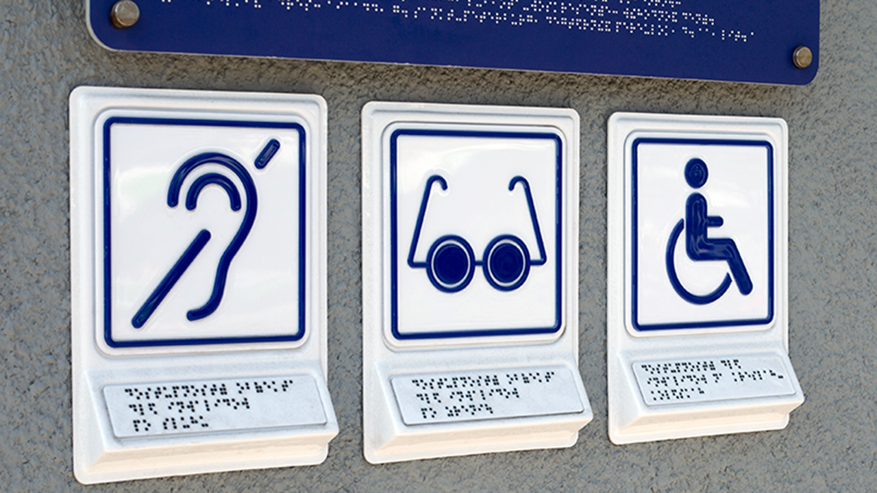 Accessibility signage in an urban environment, including the international symbols of access for hearing loss, access for low vision, and the wheelchair symbol for individuals with limited mobility.