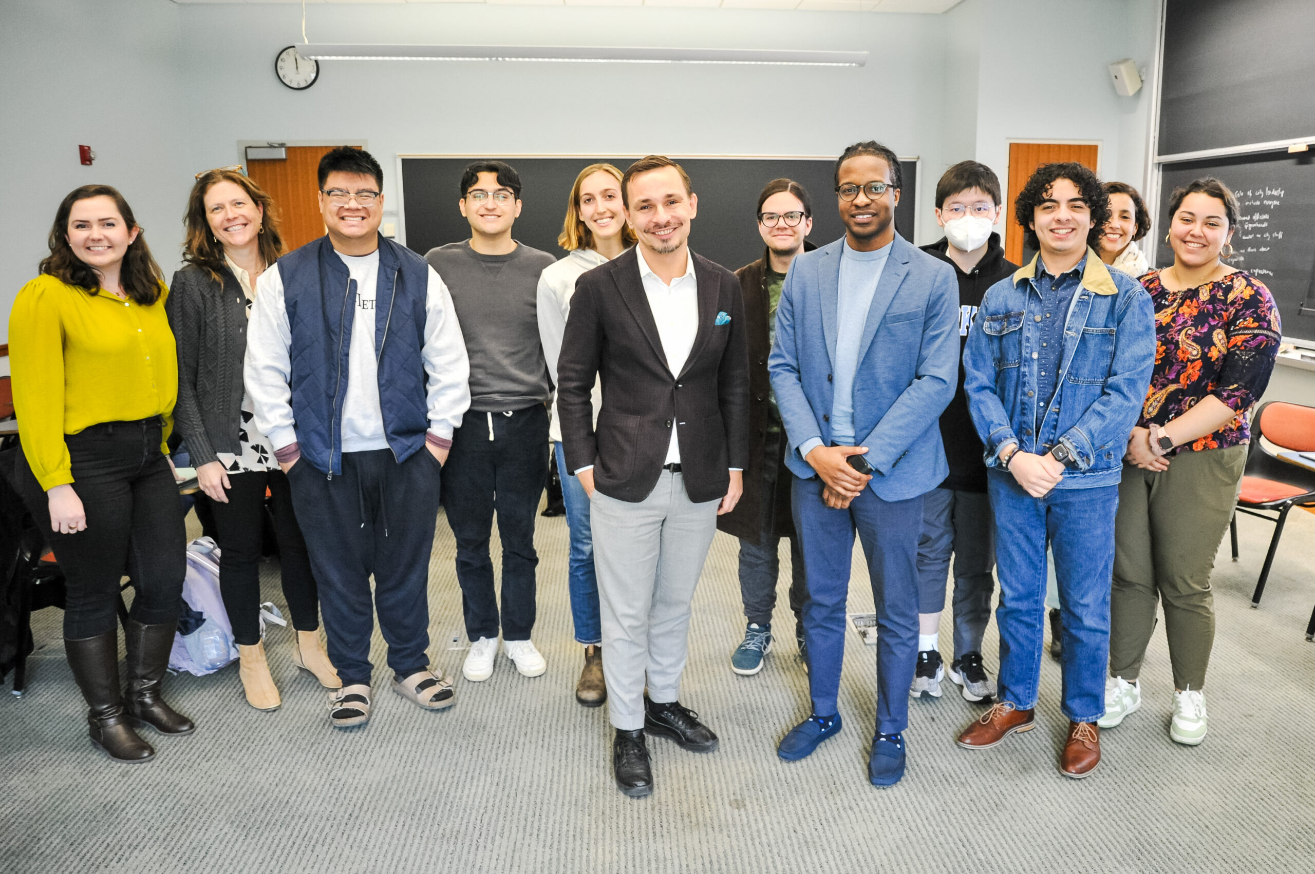 Group of students posing in a classroom and smiling at the camera, posing behind Bloomberg Public Innovation Fellows Tommi Laitio and Terrance Smith.