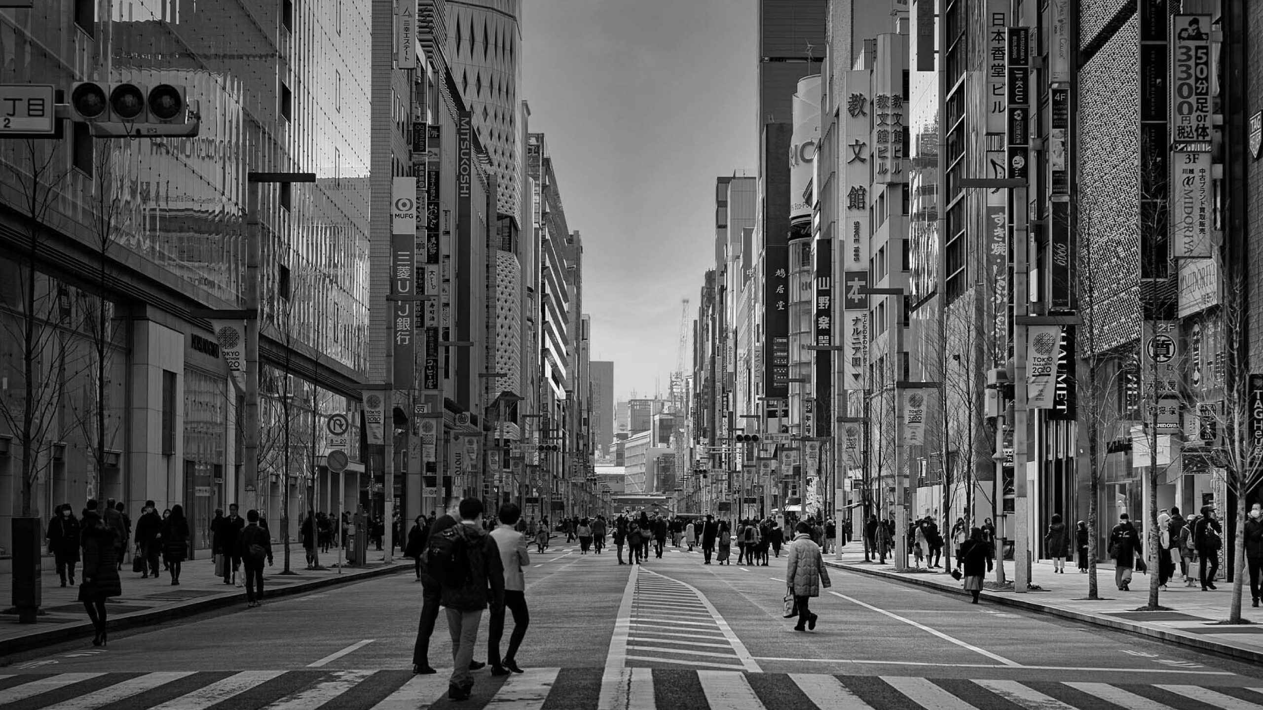 Black-and-white photograph of a bustling street in Tokyo, with pedestrians crossing a wide thoroughfare lined with numerous signs and buildings.