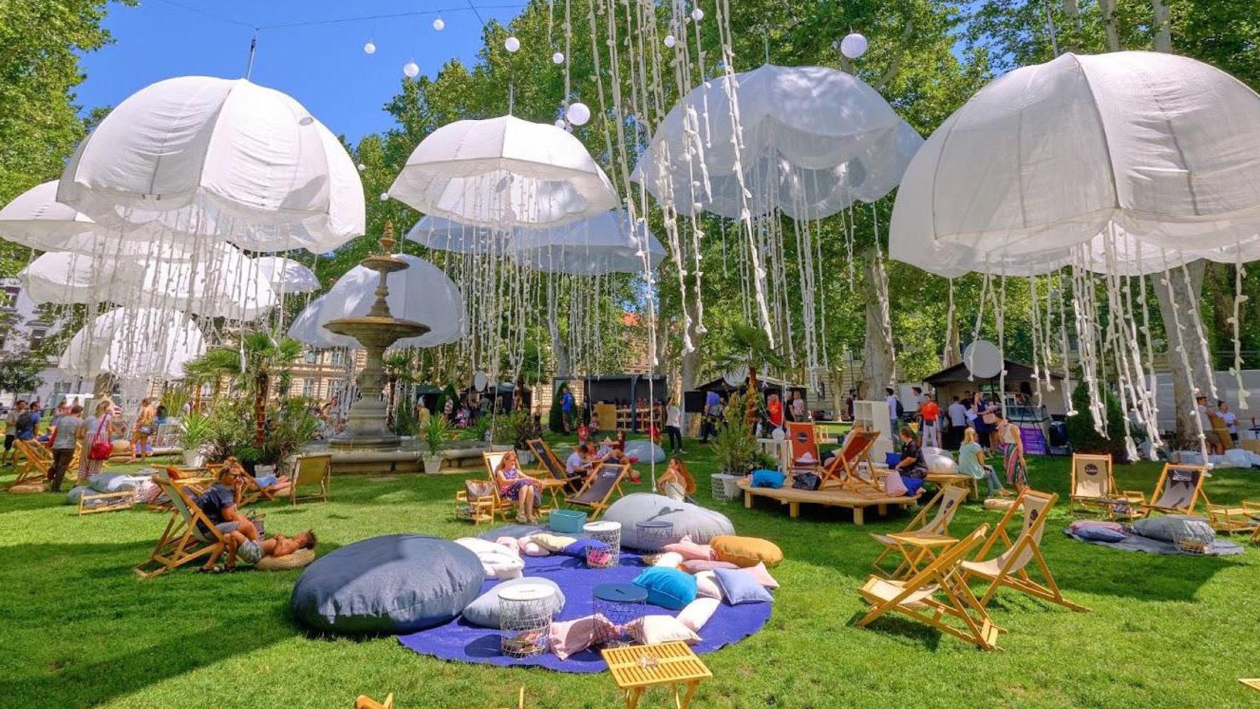 Outdoor park featuring large white umbrella sculptures hanging from trees, with colorful cushions and chairs below and visitors enjoying the ambiance.