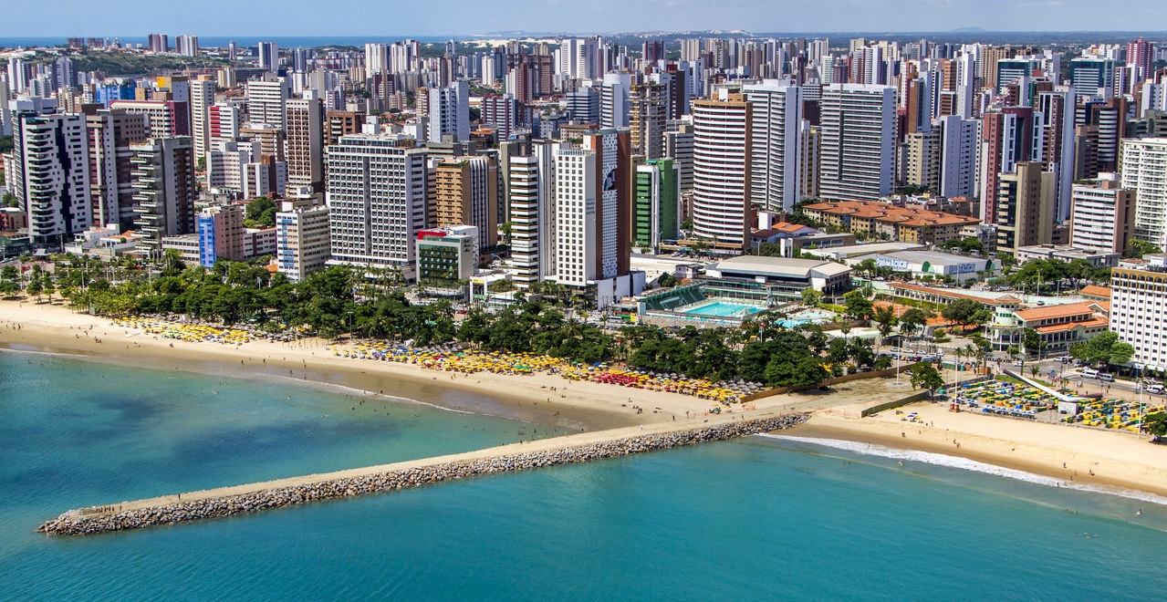 Fortaleza, Brazil Aerial view of Fortaleza's coastline showing densely packed high-rise buildings adjacent to a sunlit sandy beach and calm blue waters.