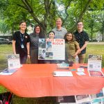 Janet Ortíz Five individuals standing behind a table with a red cloth at an outdoor event, featuring various pamphlets and a sign that reads "Love Your Block."