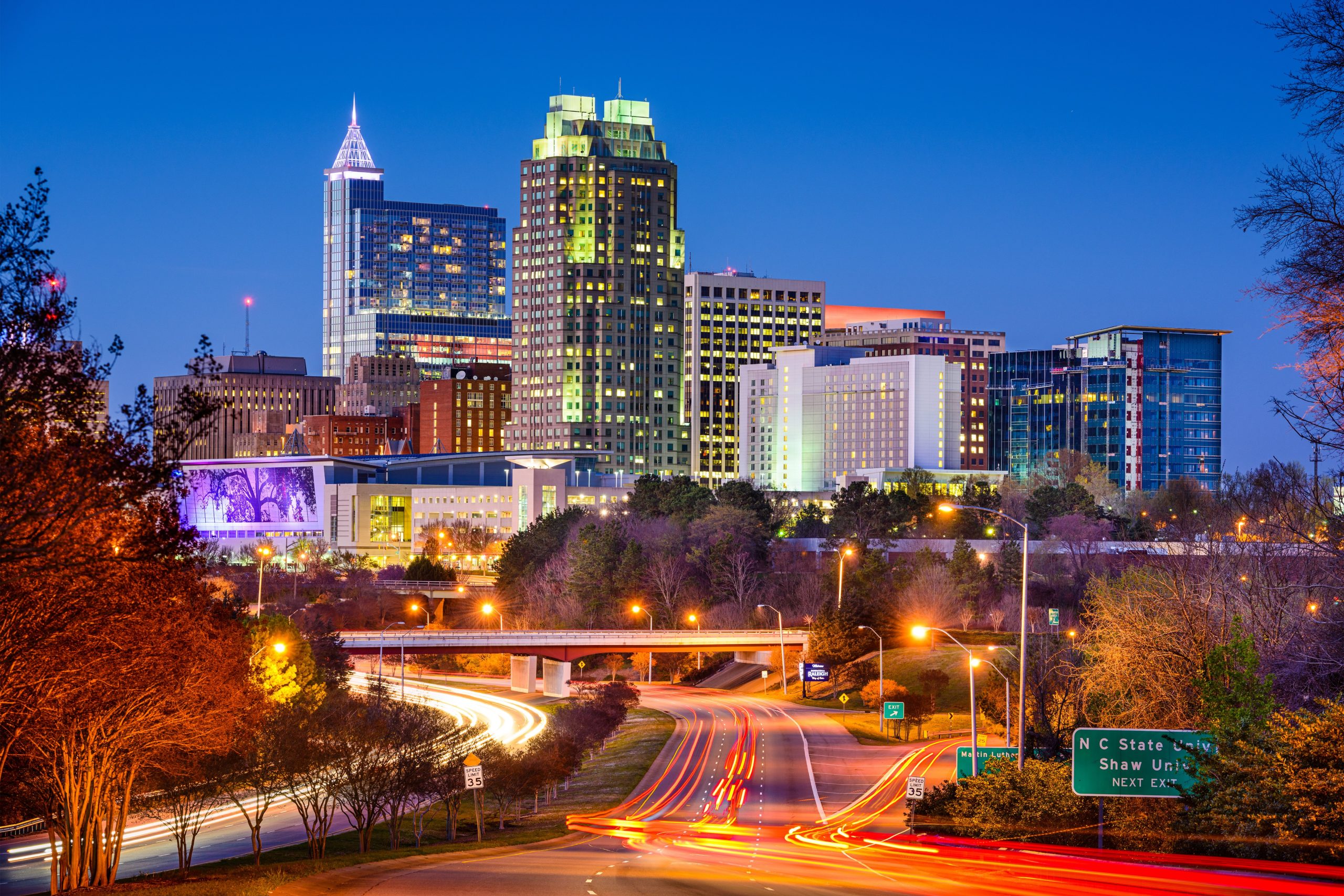 Raleigh, North Carolina Nighttime view of Raleigh, North Carolina skyline with illuminated buildings and light trails from vehicles on the highway.